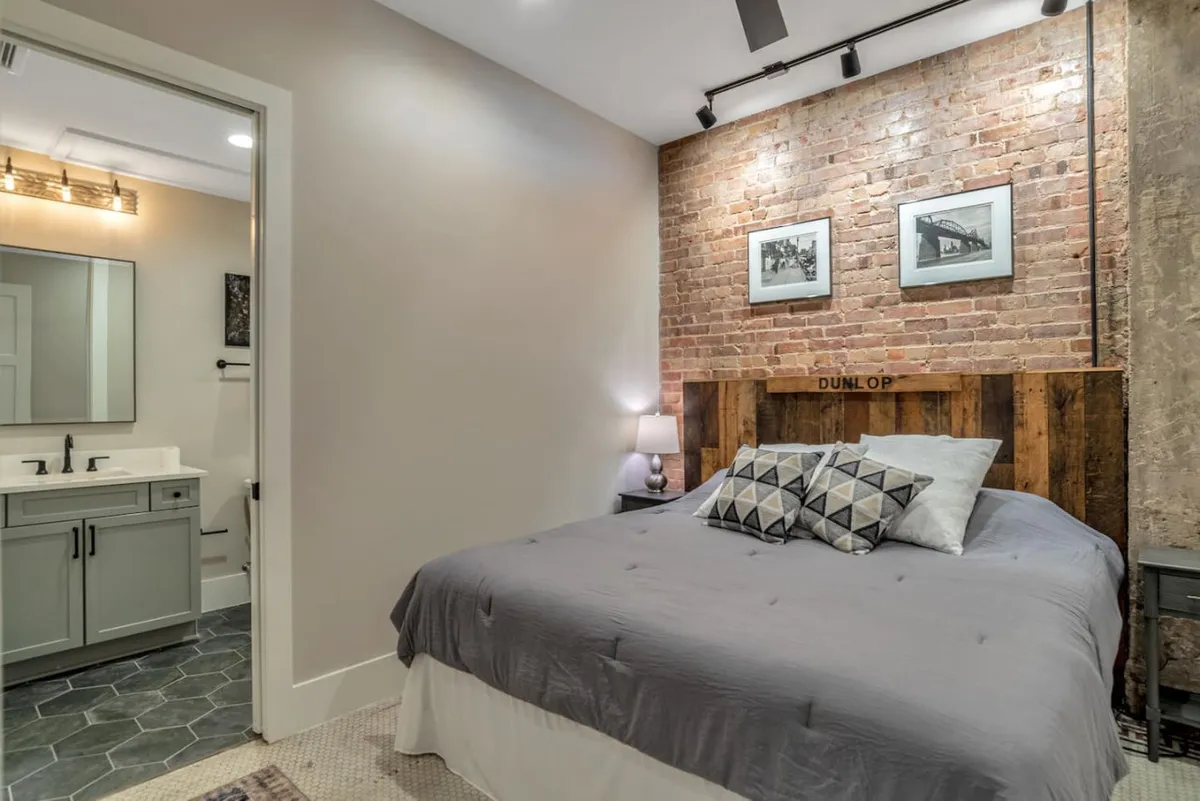 A cozy king bedroom featuring a warm exposed brick wall, a stylish wooden headboard, and decorative pillows. Soft morning light filters in, enhancing the inviting, restful mood. The room showcases plush bedding, blackout shades, and unique design details that create a private sanctuary.