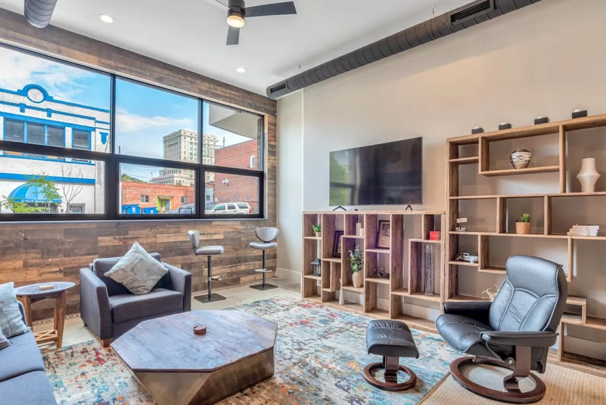The image showcases a stylish, sunlit living space with large windows that invite natural light, a modern wooden shelving unit, plush seating perfect for relaxing or socializing, a vibrant decorative rug, and a sleek TV unit. The atmosphere blends contemporary design with historic touches, radiating warmth and comfort.