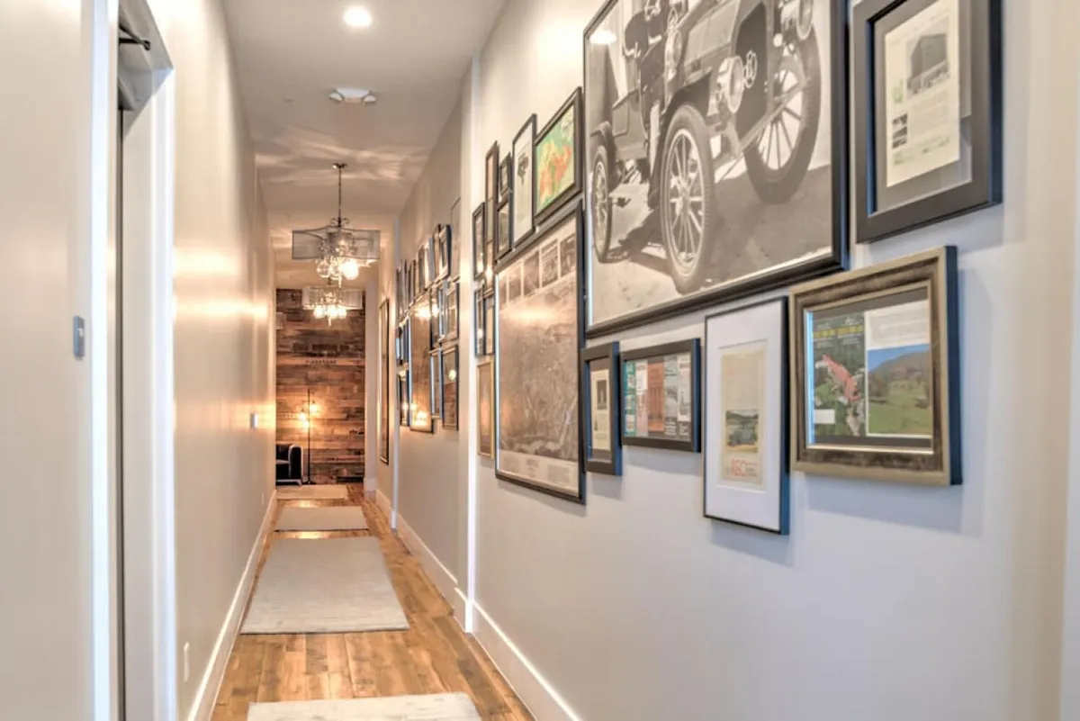 A hallway with warm wooden flooring, adorned with a gallery wall filled with framed photos and artwork. The space is illuminated by a chandelier, creating an inviting atmosphere as you transition through the entry toward the condo interior.