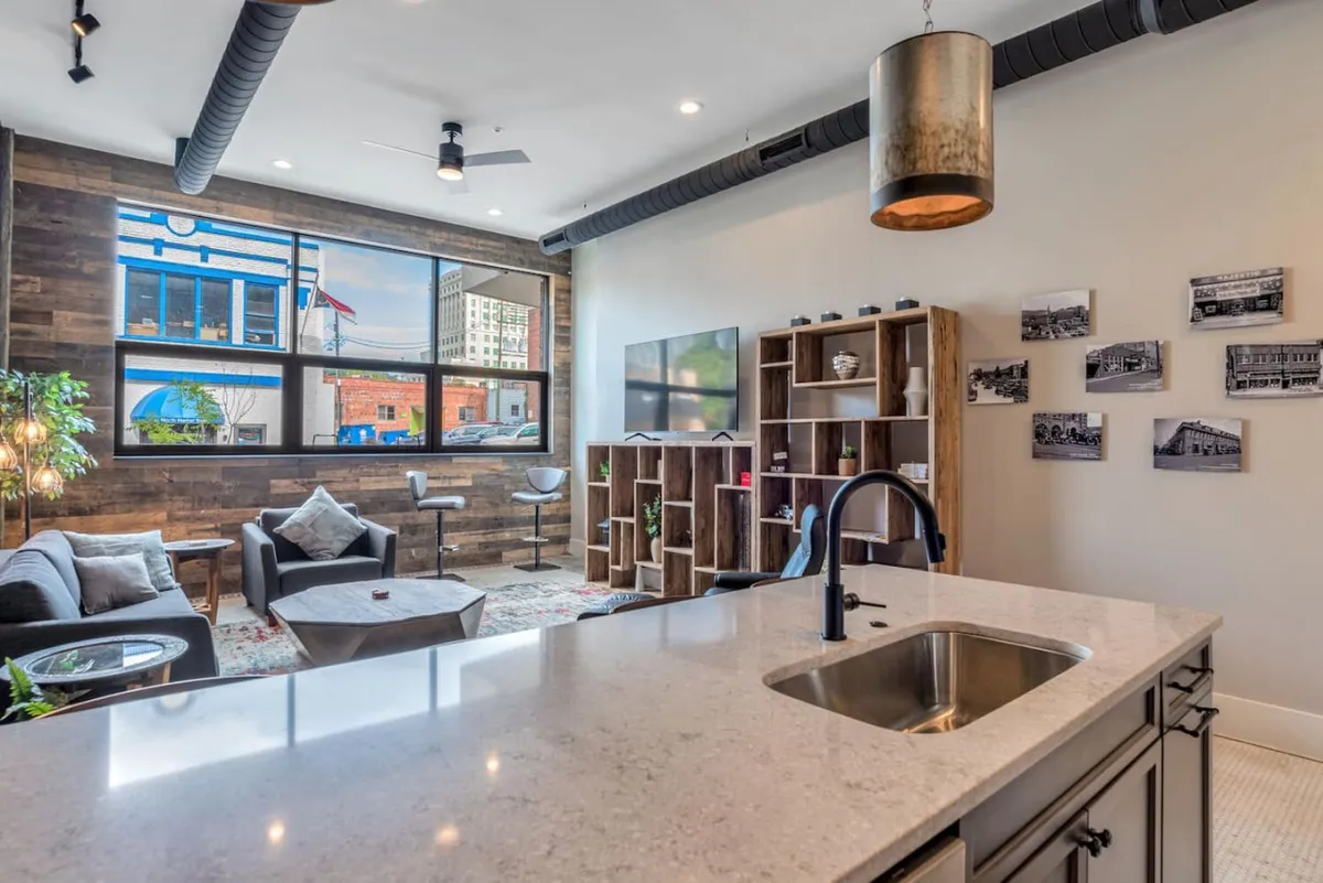 A modern, open-concept living area that seamlessly connects a comfortable seating zone with a kitchen island. Large windows offer expansive city views and let in ample sunlight, while the wooden accent wall and decorative shelving add warmth and sophistication. The space is designed for social interaction, relaxation, and enjoying the surrounding urban landscape.