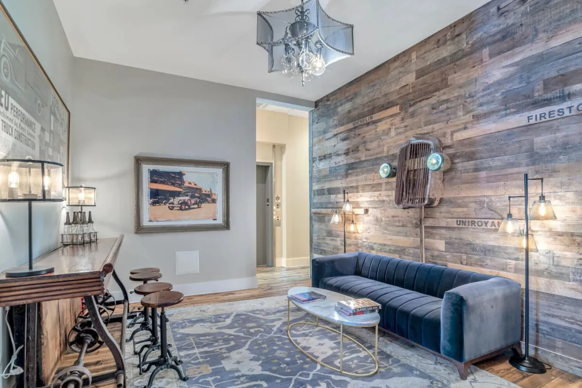 This image showcases a welcoming living area featuring a striking wooden accent wall, a plush velvet sofa, an elegant chandelier, and thoughtfully curated artwork. The overall design radiates contemporary elegance while nodding to classic charm, with high ceilings enhancing the space's grandeur. Socializing, reading, and relaxing are the main activities depicted, underscoring the space’s versatility.