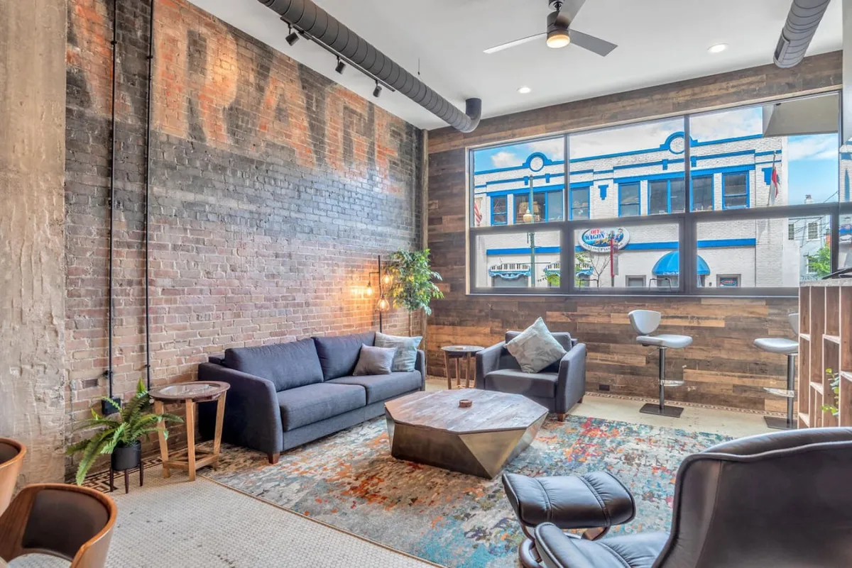 A bright, open living space featuring a comfortable sofa, modern coffee table, exposed brick wall, and a large window that floods the room with natural light. An accent chair offers additional seating, while the overall arrangement is ideal for both casual get-togethers and quiet evenings. The scene feels both cozy and contemporary, with an emphasis on natural lighting and inviting finishes.