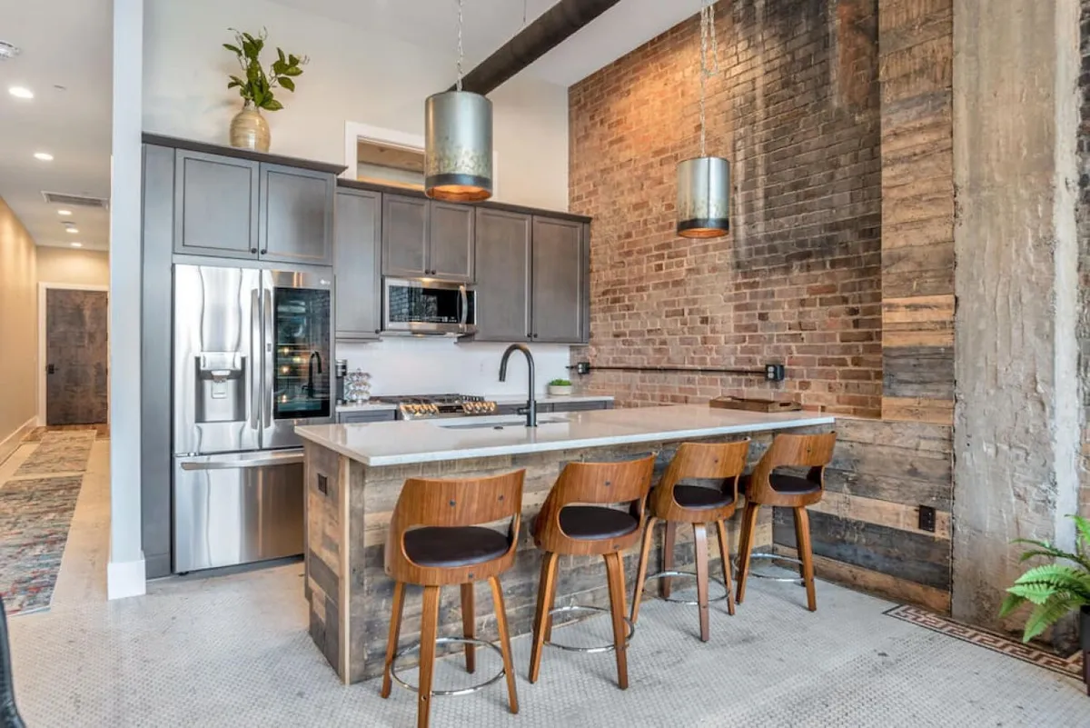 A bright, contemporary kitchen anchored by a spacious island with bar stools, set against an exposed brick wall and illuminated by elegant pendant lights. The scene is welcoming, with modern appliances visible and thoughtfully arranged for both cooking and socializing.
