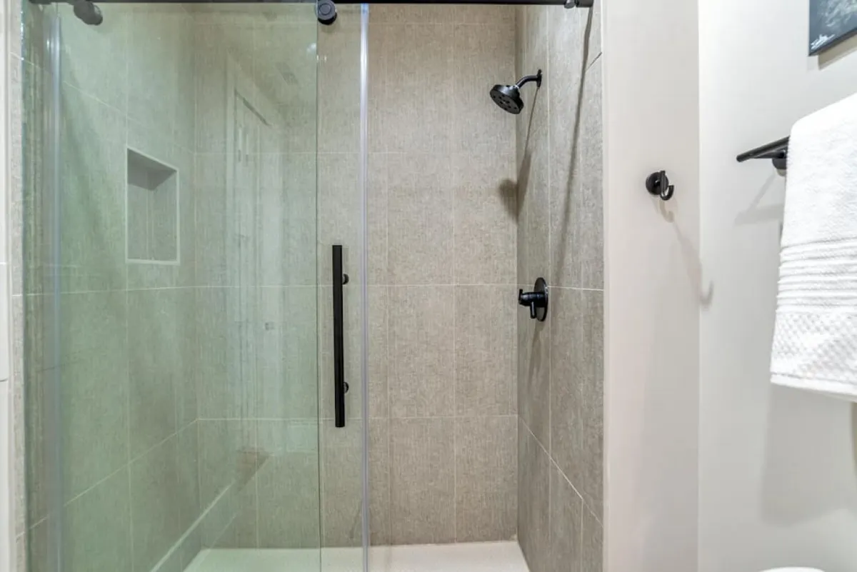 A sleek, modern bathroom with a glass shower enclosure, black fixtures, and neutral tile. The space feels spa-like and refreshing, ideal for relaxing routines. The design is clean and minimal, echoing the modern en suite bath feature.