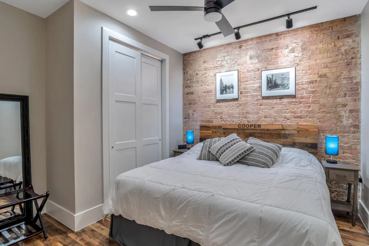 A second king bedroom with a cozy bed set against an exposed brick wall, blue lamps, and framed artwork. The space exudes tranquility, offering the perfect setting for restful sleep or leisurely mornings with a book or coffee. Ample storage and blackout shades are subtly visible.