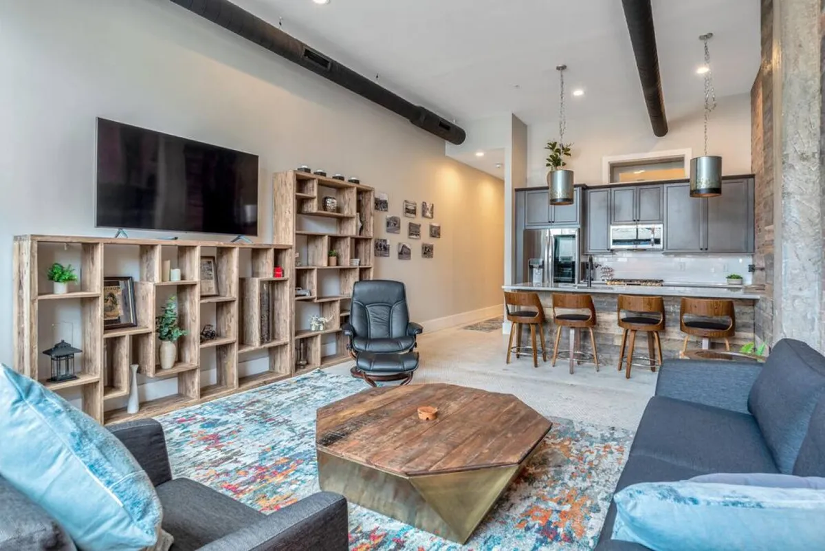 A cozy open-plan living area with a clear view into the kitchen. Decorative shelves, a TV, a wooden coffee table, and a colorful rug create a relaxed ambiance. The kitchen appears accessible and integrated into the living space, suggesting ease of movement between areas.